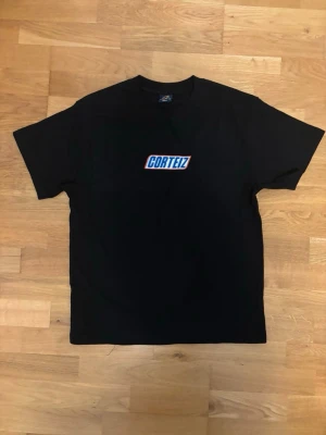 Svart Corteiz t-shirt med tryck - Svart t-shirt från Corteiz med ett färgglatt tryck på bröstet. Klassisk passform och rund halsringning. Perfekt för dig som gillar streetwear och vill ha en enkel men snygg look.
