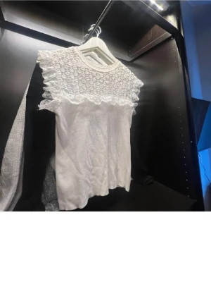 Vit blus med spets och volang - Söt vit blus med detaljerad spets upptill och volangkant längs axlar och bröst. Blusen har rund halsringning och är kortärmad. Perfekt för dig som gillar romantisk och feminin stil.