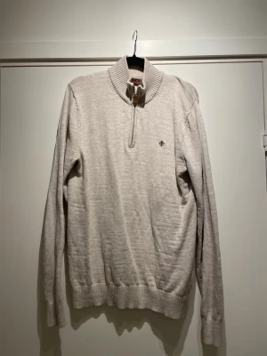 Beige stickad tröja från Morris - Snygg beige stickad tröja från Morris med halv dragkedja och ribbad krage. Tröjan har långa ärmar, ribbade muddar och en liten broderad logga på bröstet. Perfekt för lager på lager och stilrena outfits.