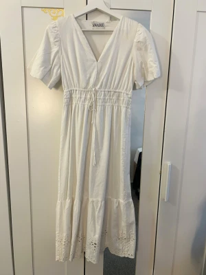 Vit långklänning med broderade detaljer - Söt vit långklänning med v-ringning, korta puffärmar och snörning i midjan. Klänningen har broderade detaljer längst ner och är tillverkad i ett mjukt bomullstyg. Perfekt för sommarens alla tillfällen!