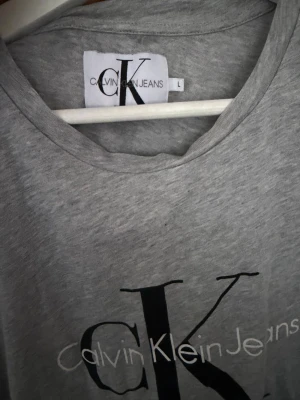 Grå Calvin Klein Jeans t-shirt - Snygg grå t-shirt från Calvin Klein Jeans med stort svart CK-tryck och vit text på bröstet. Klassisk rund halsringning och korta ärmar. Tillverkad i mjuk bomull för en skön känsla och enkel stil. Skrynklig pågrund av att den legat nervikt i en kartonglåds under längre tag. Ganska små i storleken så sitter ganska tajt 