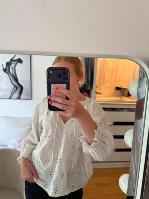 Vit blommig blus med v-ringning - Söt vit blus med v-ringning och broderade hålmönster. Blusen har långa ärmar med volangkant vid ärmslut och en avslappnad passform. Perfekt till jeans eller kjol för en trendig look.