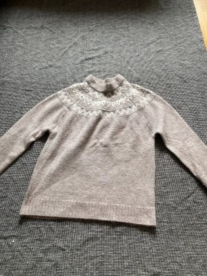 Beige stickad tröja med mönster - Mysig beige stickad tröja med ljusa mönster runt halsen. Tröjan har hög krage, långa ärmar och ribbade muddar. Använd en gång.