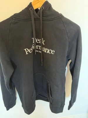 Svart hoodie Peak Performance L - Snygg svart hoodie från Peak Performance med vit logga på bröstet. Klassisk modell med huva och dragsnören samt en stor magficka framtill. Tillverkad i mjukt bomullsmaterial som är skönt att bära. Perfekt för en avslappnad och sportig stil.