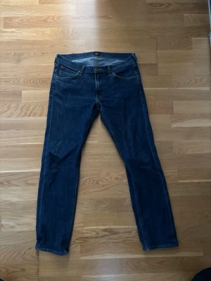 Mörkblå jeans från Lee - Säljer ett par klassiska mörkblå jeans från Lee med rak passform och fem fickor. Jeansen har gul kontrastsöm och är tillverkade i slitstarkt denimtyg. Perfekta för en avslappnad och stilren look.
