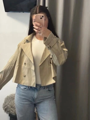 Kort beige trenchcoat med stora slag - Trendig kort trenchcoat i beige med dubbelknäppning och stora slag. Jackan har bältesdetaljer vid ärmsluten och en boxig, avslappnad passform. Perfekt för dig som gillar en modern twist på klassikern. Materialet ser ut att vara bomull eller bomullsblandning.