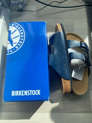 Mörkblå Birkenstock sandaler - Snygga mörkblå sandaler från Birkenstock med två justerbara remmar och metallspännen. Köpte från Monaco men dom var betydligt stora för mig. Fotbädden är i kork och sulan är svart. Perfekta för dig som vill ha både stil och komfort. Klassisk design som funkar till det mesta. Den passar bara för dom som har 45 uppåt:)