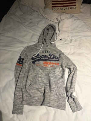 Gråmelerad hoodie från Superdry - Snygg gråmelerad hoodie från Superdry med stor logga och japansk text på bröstet. Har huva med vita dragsnören, känguruficka och tryck på ena ärmen i blått och orange. Perfekt för en avslappnad och trendig stil.