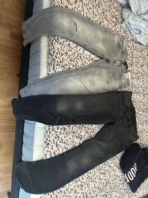 Två par slitna jeans i grått och svart - Säljer två par jeans, ett par ljusgrå med slitningar och hål samt ett par svarta med lätt tvättad look. Båda har klassisk femficksmodell och raka ben. Perfekta för dig som gillar en avslappnad och trendig stil.