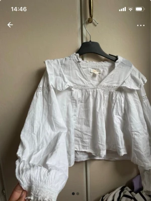Vit blus med volang från Gina Tricot - Söt vit blus från Gina Tricot med volangkrage och v-ringning. Blusen har långa puffärmar med smock vid ärmsluten och fina spetsdetaljer. Tillverkad i ett lätt bomullstyg som ger en luftig känsla.