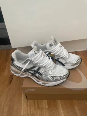 Asics Gel Kayano 14 - Snygga sneakers från Asics i vitt och silver med svarta detaljer. Skorna har en sportig design med mesh och syntetmaterial, samt tydliga Asics-loggor på sidorna. Perfekta för dig som gillar en clean och modern look.