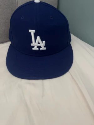 Blå LA keps från New Era - Säljer en blå 59FIFTY keps från New Era med LA-logga broderad i vitt framtill. Klassisk flat brim och strukturerad passform. Kepsen har detaljer i vitt och är tillverkad i ett slitstarkt tyg, perfekt för dig som gillar streetwear och sportig stil. In princip helt ny endast testad 2-3 ggr! Pris kan självklart diskuteras! Hör av er vid minsta fråga