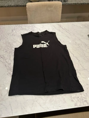 Svart ärmlös topp från Puma - Säljer en svart ärmlös topp från Puma med klassisk logga i vitt framtill. Toppen har rund halsringning och är gjord i mjuk bomull, perfekt för en sportig och avslappnad stil.