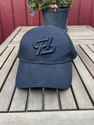 Svart meshkeps med broderad logga - Snygg svart keps med böjd skärm och mesh på sidorna för extra ventilation. Framtill sitter en stor broderad logga i svart ton-i-ton. Justerbar baktill för perfekt passform. Passar dig som gillar stilrena accessoarer med sportig känsla.