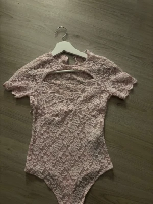 Ljusrosa spetsbody  - Säljer en ljusrosa body i spets med korta ärmar och vackert blommönster. Bodyn har en rundad halsringning och knäppning i grenen. Perfekt för dig som gillar romantisk och feminin stil.