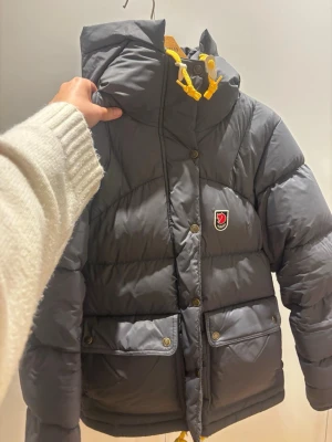 Grå pufferjacka med huva från Fjällräven - Jag säljer jackan då jag vill köpa en större storlek. Fråga jättegärna om mer bilder om ni vill och pris kan diskuteras. Snygg grå pufferjacka från Fjällräven med stor huva och gula detaljer på snörningen. Jackan har två stora fickor framtill med knappar och klassisk Fjällräven-logga på bröstet. Perfekt för kalla vinterdagar och riktigt varm.