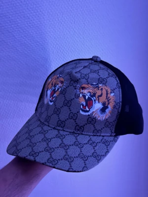 Gucci keps med tigertryck - Säljer en snygg Gucci keps med klassiskt GG-mönster och två stora tigerhuvuden framtill. Kepsen har svart mesh på sidorna och justerbar rem bak. Materialet är en mix av canvas och mesh, perfekt för dig som vill sticka ut med en unik accessoar.