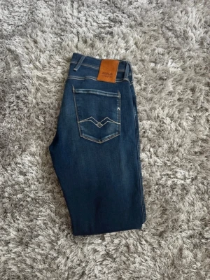 Blåa Replay Anbass - Snygga blå jeans från Replay med klassisk femficksmodell och ljusa slitningar framtill. Jeansen har en skinnpatch med logga bak i midjan och kontrastsömmar. Perfekta för en avslappnad och trendig look.