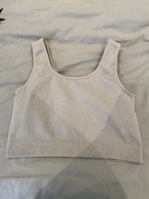 Beige ribbad crop top - Säljer en beige ribbad crop top med breda axelband och rundad halsringning. Toppen är stretchig och har en enkel, stilren design som passar perfekt till jeans eller kjol. Materialet känns mjukt och följsamt mot huden.