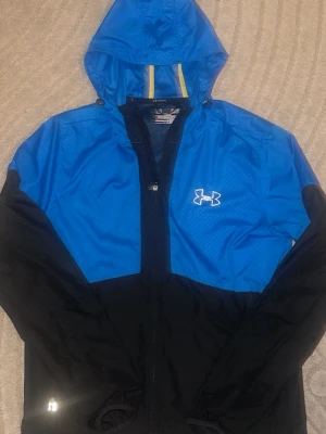 Blå och svart vindjacka Under Armour - Snygg blå och svart vindjacka från Under Armour med huva och dragkedja framtill. Jackan har meshfoder, reflekterande detaljer och logga på bröstet. Perfekt för löpning eller utomhusaktiviteter. Lätt material som andas och ger bra rörelsefrihet.