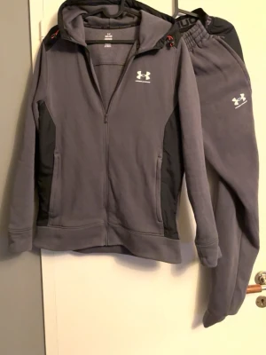 Grått träningsset från Under Armour - Säljer ett grått träningsset från Under Armour med zip-hoodie och matchande byxor. Setet har svarta detaljer på sidor och huva, samt vita loggor på både tröja och byxa. Perfekt för gymmet eller chill. Mjukt material och bekväm passform.