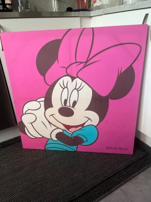 Tavla med Minnie Mouse motiv - Stor fyrkantig tavla med motiv av Minnie Mouse på rosa bakgrund. Minnie har en stor rosa rosett på huvudet och en blå tröja. Perfekt för att liva upp ett rum med färg och Disney-känsla.