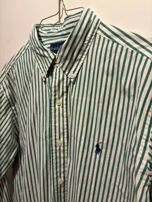 Ralph Lauren skjorta - Snygg skjorta med gröna och vita vertikala ränder. Klassisk krage, knäppning framtill och lång ärm. Liten broderad logga på bröstet. Perfekt till jeans eller chinos för en clean look.