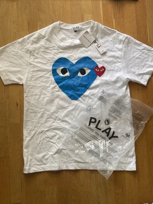 Play t shirt - Tja! Jag säljer en Snygg vit t-shirt från Comme des Garçons Play med stort blått hjärta och ett litet rött hjärta med ögon på bröstet. Klassisk passform och rund hals. Perfekt statement-plagg för dig som gillar streetwear och coola prints. Hör av er vid frågor!