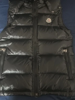 Svart dunväst från Moncler - Snygg svart dunväst från Moncler med glansig finish och klassisk logga på bröstet. Västen har två dragkedjeförsedda fickor och hög krage med tryckknappar. Perfekt för lager-på-lager och streetstyle. ANVÄND ETT PAR GÅNGER TAGS OCH PÅSEN FINNS KVAR/PRIS KAN DISKUTERAS!!!!