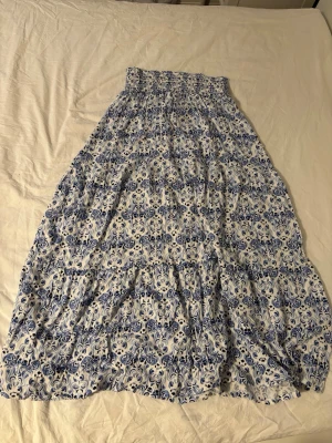 Blåvit långkjol med blommigt mönster - Supersöt långkjol i blått och vitt med blommigt mönster. Kjolen har smock i midjan för skön passform och är gjord i ett lätt och luftigt material, perfekt för varma dagar. Passar dig som gillar romantisk och bohemisk stil.