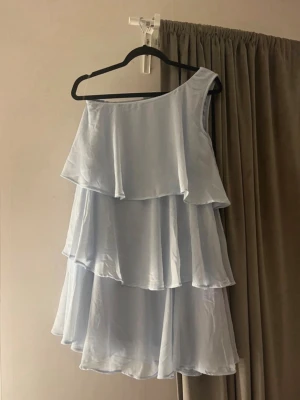 Ljusblå oneshoulder volangklänning - Säljer en ljusblå kortklänning med flera lager volanger och oneshoulder-design. Klänningen har en luftig och flowy passform, perfekt för varma dagar. Materialet känns lätt och svalt, troligen polyester eller liknande. Supersöt och trendig modell!