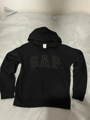 Svart hoodie från GAP Kids XL - Snygg svart hoodie från GAP Kids i storlek XL. Klassisk design med stor GAP-logga framtill och huva. Tillverkad av minst 20% återvunnet material. Perfekt för en avslappnad och cool stil.