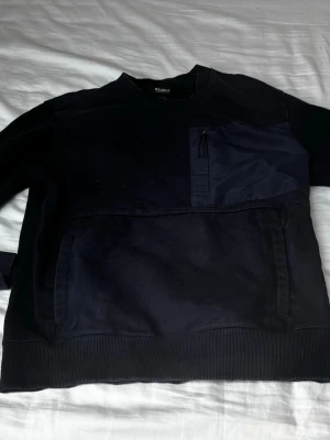 Svart sweatshirt från Pull&Bear - Cool svart sweatshirt från Pull&Bear med ribbade muddar och en bröstficka med dragkedja. Tröjan har en avslappnad passform och är gjord i mjukt material, perfekt för streetwear-stil. Praktiska fickor framtill och diskreta detaljer. Pris kan diskuteras 