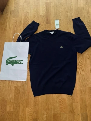 Mörkblå stickad tröja från Lacoste - Snygg mörkblå stickad tröja från Lacoste med klassisk rund halsringning och den ikoniska gröna krokodilloggan broderad på bröstet. Tröjan har långa ärmar och ribbade muddar. Perfekt för en stilren och avslappnad look. Nypris 1199kr