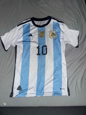 Argentina matchtröja nummer 10 - Säljer en officiell Argentina matchtröja från Adidas med klassiska blå och vita ränder. Tröjan har nummer 10 på bröstet, AFA-emblem och FIFA-märke. Svart krage och detaljer, samt broderade loggor. Perfekt för fotbollsfans!