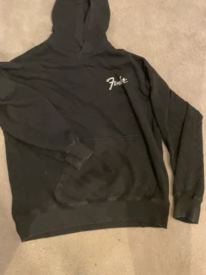 Svart Fender hoodie med tryck - Säljer en svart hoodie från Fender med vit logga på bröstet och stort tryck av en Deluxe Reverb-amp på ryggen. Klassisk känguruficka framtill och huva. Perfekt för dig som gillar musik och vill ha en skön, avslappnad stil.