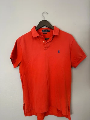 Röd/orange pikétröja från Polo Ralph Lauren - Klassisk orangeröd pikétröja från Polo Ralph Lauren med blå broderad logga på bröstet. Tröjan har krage, knappar vid halsen och korta ärmar. Perfekt till en avslappnad och stilren look.