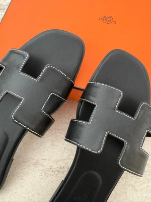 Svarta Oran sandaler i läder - Svarta Oran sandaler i slätt läder med ikonisk H-design och vita kontrastsömmar. Sandalerna har fyrkantig tå och låg sula, vilket ger en stilren och modern look. Perfekta för en trendig och avslappnad stil. Box ingår ej