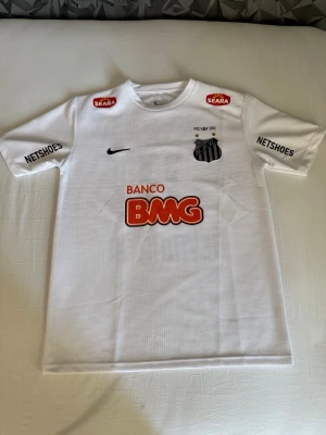 Vit Santos Neymar Jr matchtröja - Säljer en vit fotbollströja från Santos FC med Neymar Jr och nummer 11 på ryggen. Tröjan har korta ärmar, klubbmärke på bröstet, flera sponsorer och Nike-logga. Perfekt för dig som älskar brasiliansk fotboll och vill ha en ikonisk supportertröja.