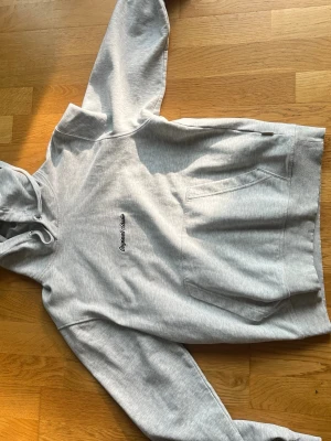 Ljusgrå hoodie med broderad text - Snygg ljusgrå hoodie med broderad text 'Originals Studio' på bröstet. Klassisk känguruficka framtill och justerbar huva med dragsko. Tillverkad i mjukt sweatshirtmaterial som är perfekt för chill dagar.