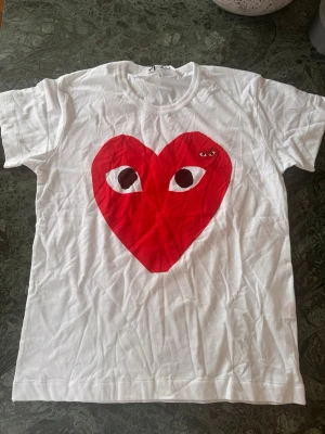 Vit Comme des Garçons PLAY t-shirt - Säljer en vit t-shirt från Comme des Garçons PLAY med det ikoniska röda hjärtat med ögon på bröstet. T-shirten har rund hals och korta ärmar, tillverkad i mjuk bomull. Perfekt för dig som gillar streetwear och vill sticka ut.