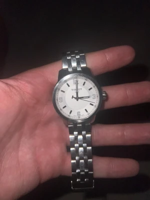 Tissot klocka med silverlänk - Stilren Tissot klocka med rund vit urtavla och tydliga svarta markeringar. Klockan har en klassisk länk i silverfärgat metall och enkel, tidlös design. Perfekt accessoar för dig som gillar en clean look. Klockan behöver byta batteri samt storlek beroende på om den passar eller inte därför säljer jag den för en billigare peng en vanligt 