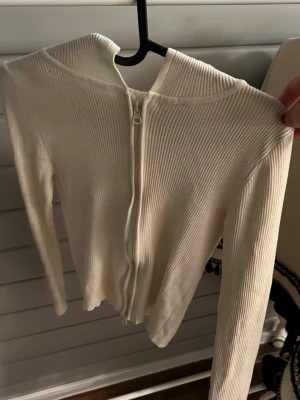Vit ribbad hoodie med dragkedja - Säljer en stilren vit hoodie i ribbat material med dragkedja framtill. Hoodien har långa ärmar och en klassisk huva. Perfekt för dig som gillar en enkel men trendig look. Materialet är stretchigt och följsamt.
