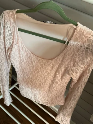Ljusrosa spetsblus med lång ärm - Söt ljusrosa blus i spets med långärmad design och rundad urringning. Toppen är croppad och har transparenta ärmar i samma spetsmaterial. Perfekt för dig som gillar romantisk och feminin stil.