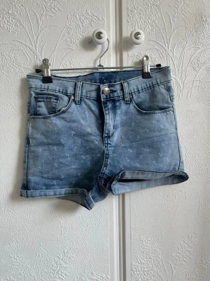 Blå jeansshorts med ankare från Uniq Young - Säljer ett par ljusblå jeansshorts från Uniq Young med små vita ankare som mönster. Shortsen har klassisk uppvikta benslut och bälteshällor. Perfekta för sommardagar och ger en chill vibe till din outfit.