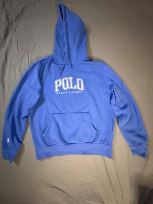 Blå hoodie från Polo Ralph Lauren - Snygg blå hoodie från Polo Ralph Lauren med stor vit logga på bröstet och klassisk polospelare broderad på ärmen. Hoodien har huva med dragsko och en stor magficka framtill. Perfekt för en chill och avslappnad stil. Detta är en äldre model som ej går o köpa längre