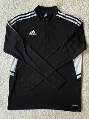 Svart Adidas träningströja med dragkedja - Snygg svart långärmad träningströja från Adidas med vita detaljer och dragkedja vid halsen. Tröjan har klassiska Adidas-ränder på axlarna och är tillverkad i funktionsmaterialet AEROREADY som transporterar bort fukt. Perfekt för träning eller uppvärmning.