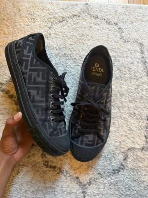 Svarta sneakers med Fendi-logga - Snygga svarta sneakers från Fendi med klassiskt FF-monogram i grått över hela skon. Skorna har svarta snören, metallöljetter och en robust gummisula. Perfekta för dig som vill ha en exklusiv och trendig look med tydlig logga och streetwear-vibe.