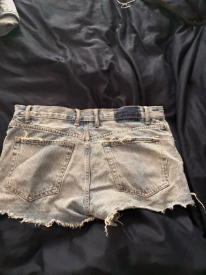 Blå jeansshorts från ZARA - Säljer ett par blå jeansshorts från ZARA med råa, fransiga kanter och klassisk femficksdesign. Shortsen har bälteshällor och slitna detaljer för en avslappnad look. Perfekta för varma dagar och enkel att matcha med allt.