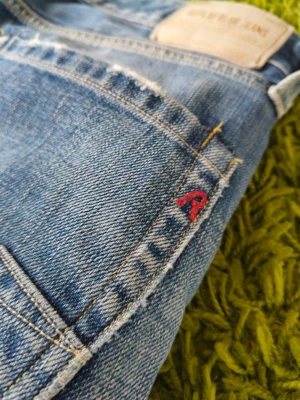 Blå raka replay jeans med röd detalj w30 l32 - Säljer ett par klassiska blå jeans med raka ben och slitna detaljer. Jeansen har bakfickor med dekorativa sömmar och en liten röd broderad detalj vid fickan. Tillverkade i denim med kontrastsömmar och patch bak i midjan.